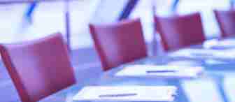 A glass conference table is set with clipboards, pens, and papers. Red chairs are arranged around the table in the background, with a soft focus on the modern, light-filled space. The overall tone of the image is cool, with blue and purple hues.