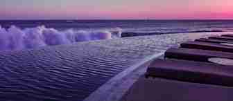 Infinity pool overlooking the ocean at sunset. Waves crash gently on the edge, and the sky is painted in hues of pink and purple. Several poolside lounge chairs are lined up on the right, suggesting a serene and luxurious setting.