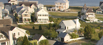 A coastal neighborhood with several large, light-colored houses near a body of water. The homes have various architectural designs, with porches and large windows, surrounded by greenery and white fences. A boat is seen in the water in the distance.