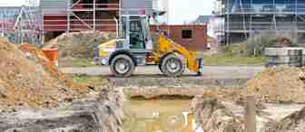 A construction site with two partially built houses in the background. In the foreground, there is a small excavator parked beside a rectangular pit filled with water. The area is surrounded by dirt mounds and construction materials.