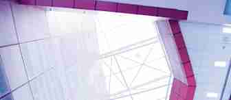 A view looking up at the geometric ceiling of a modern building, featuring large, angled glass panels that allow natural light to flood the interior. The surrounding walls are clad in red and white tiles, creating a colorful, contemporary architectural design.