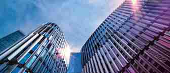 A low-angle view of modern office buildings with reflective glass and metal facades, rising towards a bright sky. The sun is shining brightly, creating lens flare effects on the buildings. The architectural structures are sleek and contemporary.
