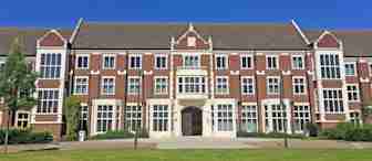 A large brick building with a symmetrical design, featuring multiple windows and a central entrance, sits against a clear blue sky. The foreground includes a well-maintained green lawn and trees on either side.