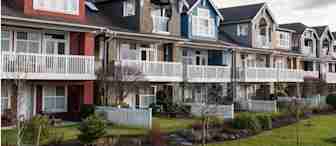 A row of stylish, multi-colored houses with large windows, spacious balconies, and gabled roofs, all lined up side by side. Each house has a small front yard with a white picket fence, and bare trees and cloudy sky can be seen in the background.