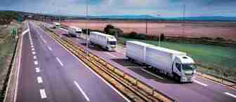 A fleet of white semi-trucks drives on a multi-lane highway through a rural area. The sky is overcast, and fields stretch out on either side of the road. Street lights line the highway, and distant mountains are visible on the horizon.