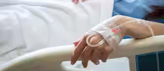 A person lies in a hospital bed, covered with a white blanket. Their hand, with an intravenous (IV) tube inserted and secured by transparent tape, rests on the bed railing. They are wearing a blue hospital gown, partially visible in the image.