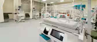 A neonatal intensive care unit with multiple incubators arranged in a spacious, well-lit room. The incubators are equipped with monitors, and chairs are placed nearby. Walls are light-colored, and the floor is clean and uncluttered.
