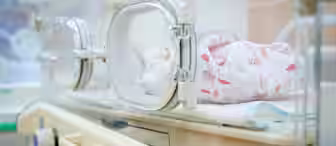 A newborn baby is peacefully sleeping inside a hospital incubator. The baby is wrapped in a patterned blanket featuring small, colorful designs. The incubator is equipped with controls and clear panels for visibility and protection.