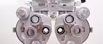 Close-up of an optometry phoropter, a device with multiple lens dials and knobs used for measuring vision. The metallic and white device is positioned against a neutral background, highlighting its intricate components.