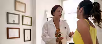 A doctor in a white coat is speaking with a patient dressed in a yellow outfit in a medical office. The doctor is holding a sphygmomanometer. Certificates are hung on the wall behind them, and medical equipment is visible in the background.