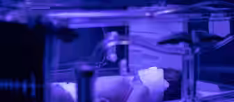 A newborn baby lies in an incubator under blue phototherapy light, wearing an eye shield for protection. The scene appears to be in a hospital neonatal unit.