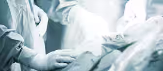 Three healthcare professionals in surgical gowns and gloves perform a medical procedure in a sterile environment. Their hands are shown in focus, working meticulously on a patient, highlighting the precision and care involved in surgery.