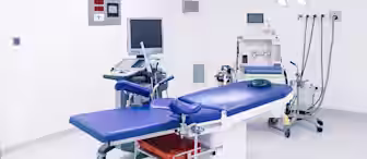 A clean, modern operating room with a blue surgical table, overhead lights, medical monitors, and various equipment against white walls and floors.