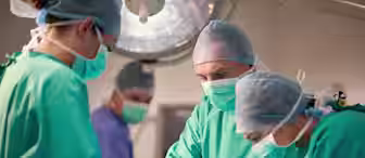 Three surgeons in green surgical gowns and masks perform an operation under a large surgical light. Another medical professional in purple scrubs stands in the background, observing. The setting appears to be a sterile operating room.