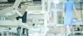 Close-up of a hospital bed's wheels on a polished floor. A medical professional in blue scrubs is seen walking away in the background, with blurred hospital equipment and lights creating a clinical atmosphere.