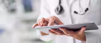 A healthcare professional wearing a white lab coat and a stethoscope around their neck is using a tablet. The background is blurred, focusing on the person's hands interacting with the screen.