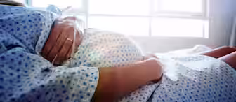 A pregnant person lying in a hospital bed wearing a hospital gown, with hands resting on their belly, sunlight streaming through a window in the background.