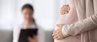 A pregnant woman gently holds her belly while standing in the foreground. In the blurred background, a healthcare professional sits and writes on a clipboard.