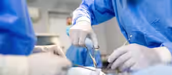 Surgeons, dressed in blue surgical gowns and gloves, perform an operation. One surgeon holds forceps, while another assists. The sterile environment and focused attention on the task are evident, highlighting the precision and care involved in the procedure.
