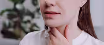 A woman in a white shirt touches her throat with one hand, suggesting discomfort or a sore throat. The background is blurred, with a green plant visible.