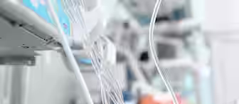 A hospital room with a blurred patient lying in bed in the background. Various medical equipment and monitoring devices, including IV lines and tubes, are focused in the foreground. The scene suggests a critical care or intensive care situation.