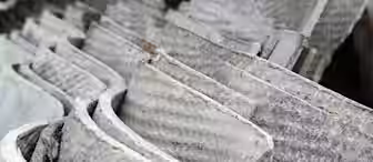 Stacked corrugated sheets of weathered, gray asbestos roofing material, showing a rough texture with visible fibers. The sheets are densely packed, illustrating an industrial or construction setting.