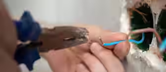 Close-up of a person using pliers to strip the insulation off a blue electrical wire. The person is installing or repairing wiring inside a wall, with a green electrical box visible in the background. The wall has some exposed wires and unfinished edges.