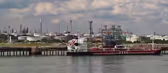 A large industrial port area with a docked cargo ship, surrounded by storage tanks and refinery structures. The water in the foreground is calm, and the background features numerous smokestacks and industrial buildings under a partly cloudy sky.