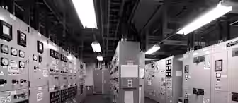 A monochrome image of an industrial control room. The room is filled with large electrical panels, meters, switches, and labels. Overhead fluorescent lights illuminate the space, casting shadows on the floor.