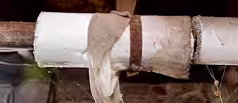 A close-up of an old, rusted pipe wrapped in white insulation and a frayed cloth, with cobwebs hanging nearby and a rough brick wall in the background.