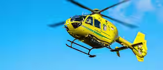 A yellow helicopter with green stripes flies across a clear blue sky. Its rotor blades are spinning, and there is a logo on the side. The helicopter is in motion, and the sunlight highlights its bright color.