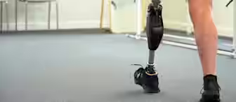 A person with a prosthetic leg, wearing black shorts and black sneakers, walks indoors on a carpeted floor; two empty chairs are visible in the background.