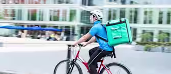 A delivery person rides a red bicycle on a city street, wearing a blue shirt, black pants, a white helmet, and carrying a large green insulated backpack. Blurred buildings and outdoor seating areas are visible in the background.