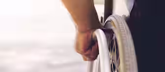 Close-up of a person’s hand gripping the wheel of a wheelchair. The scene appears to be outdoors with sunlight casting a warm glow. The wheelchair and hand are in sharp focus, while the background is blurred.