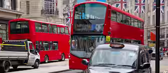 A busy urban street scene featuring two red double-decker buses, a traditional black taxi, and a service truck. The background showcases buildings adorned with British flags, indicating a vibrant city atmosphere.