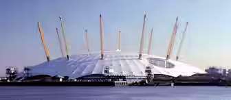 The image shows the O2 Arena, a large, white dome-shaped structure with yellow support masts, set against a clear blue sky. The structure is located by the water, with its reflection visible on the surface. Buildings can be seen in the background.