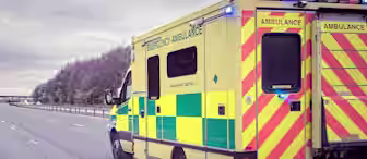 A yellow and green ambulance with "Emergency Ambulance" markings is driving on a mostly empty highway. The vehicle's rear has red and yellow chevron stripes, and the sky is overcast. There are trees and a small overpass in the distance.
