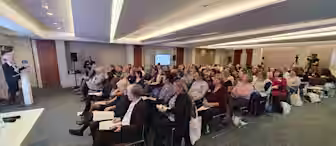 A large group of people sit facing a podium where a speaker is presenting in a modern conference room. The audience members, many holding papers or bags, are attentively listening. A screen in the background displays presentation content.