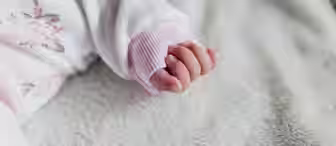 Close-up of a baby's hand resting on a soft, white textured surface. The baby is wearing a long-sleeved, white garment with pink floral patterns and light pink cuffs. The fingers are gently curled, and the overall scene conveys a sense of calm and softness.