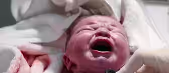 A newborn baby with a reddish complexion is crying loudly while lying on a white towel. A gloved hand is holding a stethoscope to the baby's chest, performing a health examination. The baby is wrapped in a white cloth.