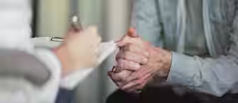 A person holding a pen and clipboard sits across from an elderly man who is resting his clasped hands on his knees. The elderly man wears a light grey shirt and looks contemplative. The scene appears to be a discussion, possibly in a medical or counseling setting.
