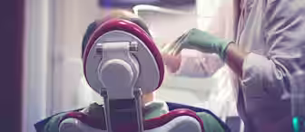 A dentist wearing a mask and gloves is treating a patient reclining in a dental chair. The dentist is focused on the patient's mouth, while various dental equipment and a monitor are visible in the background. The image captures a routine dental check-up.