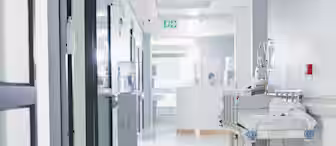 A hospital hallway with bright lighting. A medical gurney is parked against the wall, and a water cooler is nearby. In the background, a healthcare professional is visible at a reception desk. The corridor appears clean and sterile.