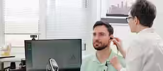 A healthcare professional uses specialized diagnostic equipment to examine a seated man's ear and throat in a medical office. A computer monitor with diagnostic graphs and other medical tools are visible on the desk.