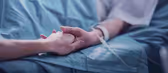 A close-up of two people holding hands. One is a patient lying in a hospital bed with an IV in their arm, while the other appears to be offering comfort and support. The scene conveys compassion and care in a medical setting.