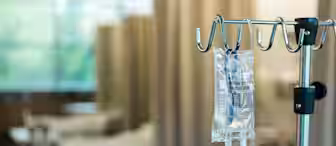 An IV drip bag hangs from a stand in a hospital room. The background shows several empty hospital beds with beige curtains separating them. The scene is softly lit, suggesting a calm, clinical environment.