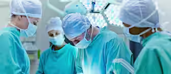 A group of surgeons and medical staff in sterile green scrubs and masks perform surgery in an operating room. The surgeons, focused on the procedure, work under bright surgical lights. One person monitors anesthesia equipment while others assist in the operation.