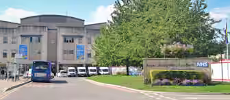A hospital entrance with a sign reading "Royal United Hospitals Bath NHS." There’s a bus at a bus stop in front, several parked vans, and tall banners featuring a rainbow. A tree with flowers and some greenery are on the right side of the sign.