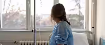 A person with long hair sits on a hospital bed wearing a medical gown, facing a window with IV drip connected to their arm. The room is softly lit, and the window reveals a view of trees and a pale sky.
