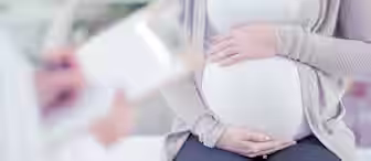 A pregnant person sits on an examination table, holding their belly. They are wearing a white shirt, a beige cardigan, and dark pants. In the foreground, a healthcare professional is taking notes on a clipboard. The setting appears to be a medical office.